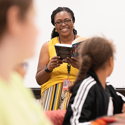 female faculty member wearing yellow reading from a book in front of class